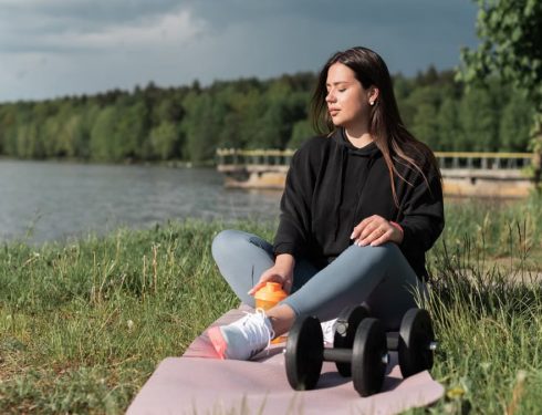Woman sitting on yoga mat beside a lake with exercise equipment, enjoying a calm outdoor workout.